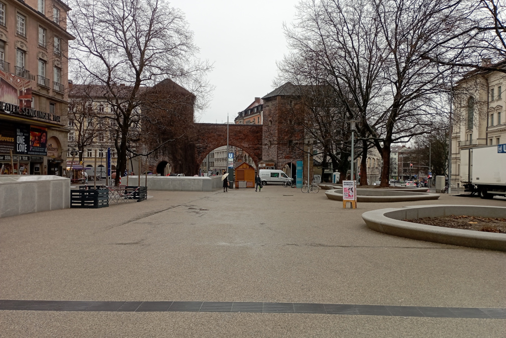 Sendlinger-Tor-Platz, Blickrichtung Sendlinger Tor. Bei Regen bietet sich dem Photographen eine graue, rau asphaltierte Fläche. Rechts große runde Tröge, auf denen man sitzen kann, mit Bäumen.